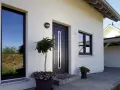 Nowoczesny dom z dużymi oknami i ciemnym **holl**em. Przed wejściem rosną drzewka i kwiaty.