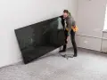 Mężczyzna z pasem narzędziowym przy ścianie trzyma duży, czarny telewizor. Zastanawia się, na jakiej wysokości telewizor będzie najlepiej wyglądał.