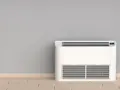 Nowoczesny klimatyzator bez jednostki zewnętrznej, stojący na drewnianej podłodze przy szarej ścianie.