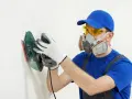 Pracownik w masce i okularach ochronnych wykonuje szlifowanie gładzi na białej ścianie za pomocą szlifierki.
