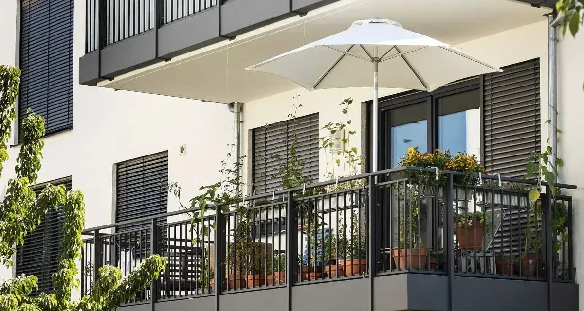 Nowoczesny balkon po remoncie, z białym parasolem i doniczkami pełnymi zieleni.