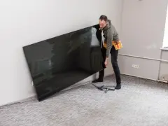 Mężczyzna z pasem narzędziowym przy ścianie trzyma duży, czarny telewizor. Zastanawia się, na jakiej wysokości telewizor będzie najlepiej wyglądał.