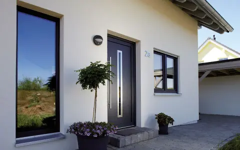 Nowoczesny dom z dużymi oknami i ciemnym **holl**em. Przed wejściem rosną drzewka i kwiaty.