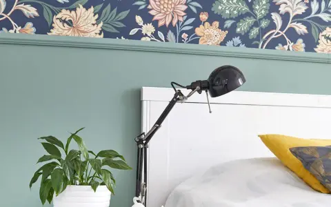 Zielona lamperia pod kwiecistną tapetą, obok łóżka stoi lampka i doniczka z rośliną.