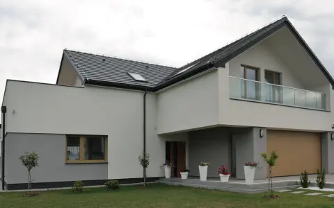 Nowoczesna elewacja domu z szarymi i białymi tynkami, dużym garażem i balkonem z przeszkloną balustradą.