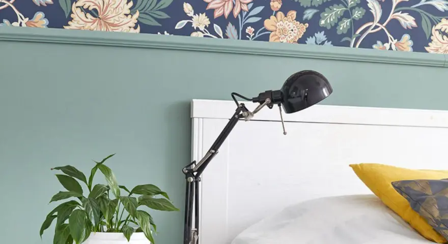 Zielona lamperia pod kwiecistną tapetą, obok łóżka stoi lampka i doniczka z rośliną.