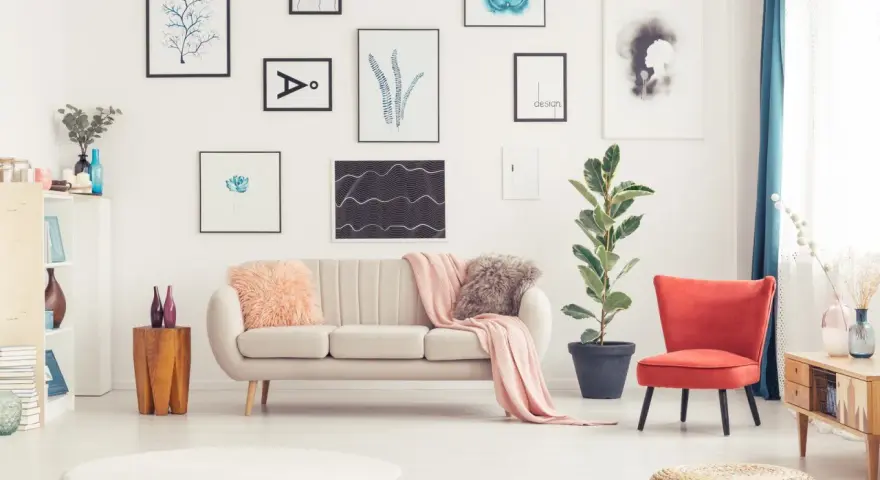 Sofa w kolorze ecru jaki to kolor? Delikatna, przytulna, idealna do salonu. Obok niej czerwony fotel i mnóstwo obrazów na ścianie.