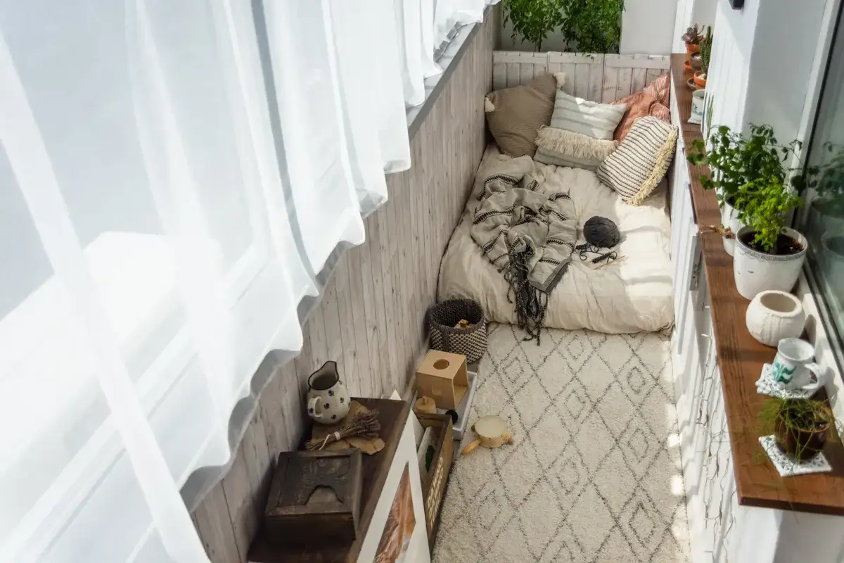 Przytulna loggia z materacem, poduszkami i kocem. Obok stolik z dzbankiem i ozdobami. Balkon zamieniony w relaksującą przestrzeń.