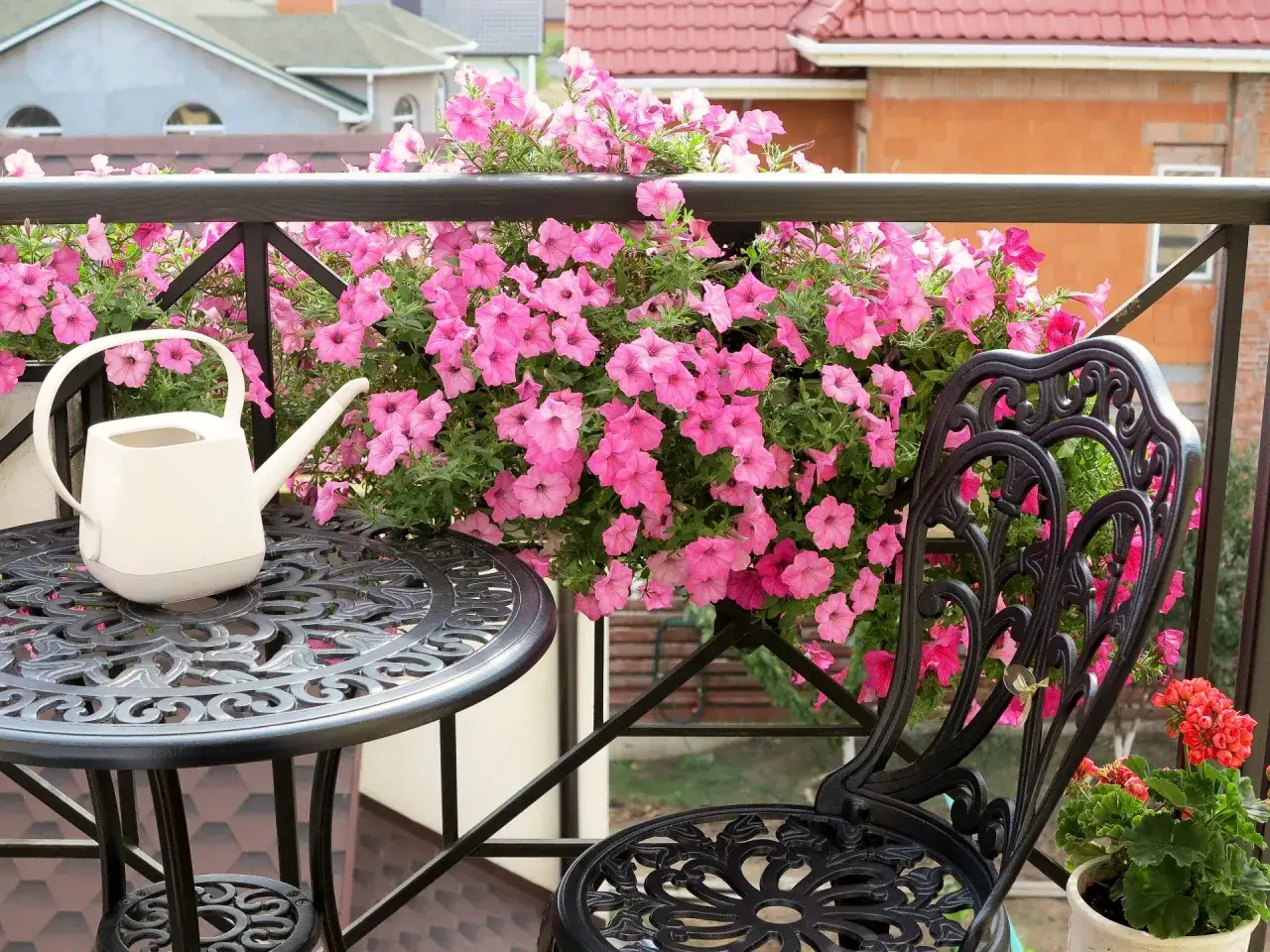 Balkon pełen różowych kwiatów, które zachwycają swoimi nazwami. Obok stolik z konewką i ozdobne krzesło.
