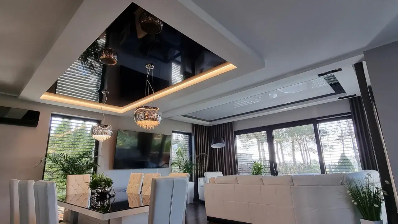 Nowoczesny salon z czarnym, błyszczącym sufitem napinanym, który odbija eleganckie lampy i okna.